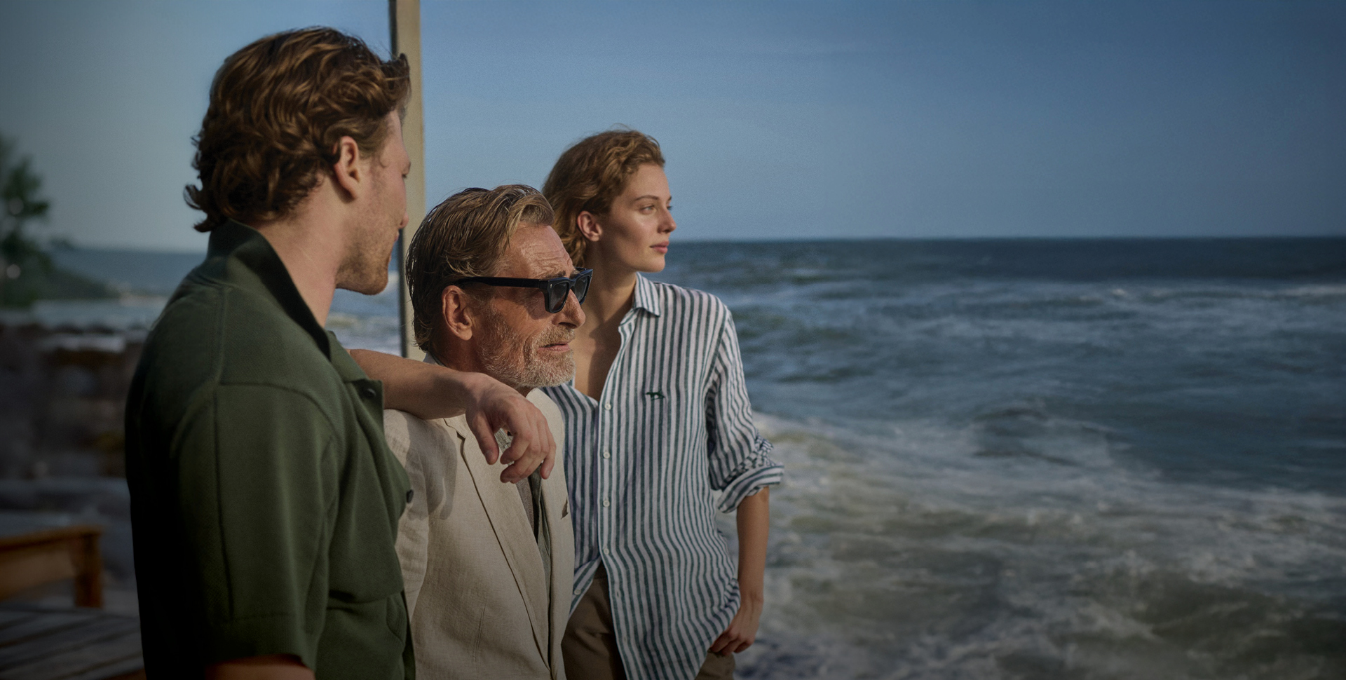 Three people standing on a seaside deck beside choppy waves, wearing an olive short-sleeve shirt, a beige blazer, and a blue-and-white striped button-down